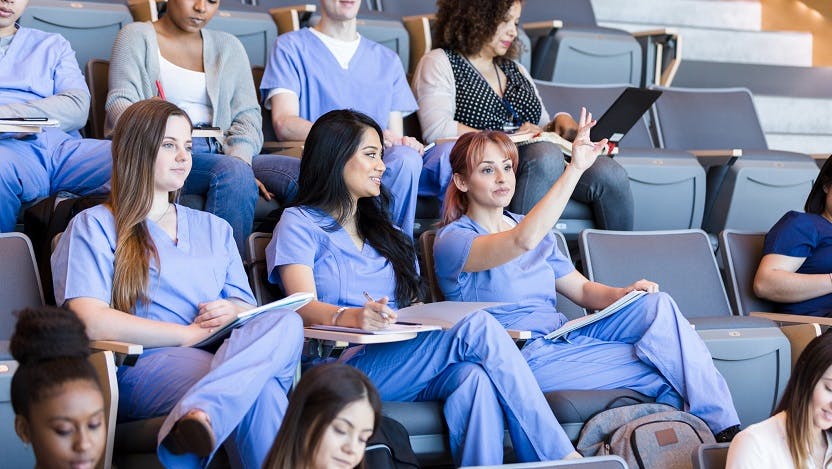 medical students wearing blue scrubs seated in a lecture hall. one is raising her hand.