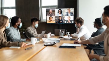 people around a table wearing masks, participating in a meeting with others online