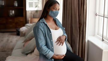Masked pregnant person at home looking out the window