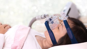 Happy and healthy senior woman sleeping deeply on her back with CPAP machine mask and hose.