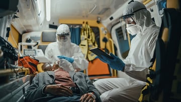 Paramedic in coveralls in an ambulance using tablet while tending for a patient on the way to hospital.