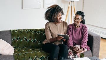 An adult sitting with a senior on a couch displaying something on a tablet