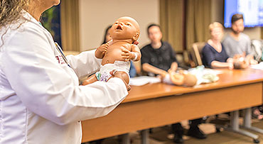 Nurse educator holds up an infant care demo doll in front of couples at a prenatal class