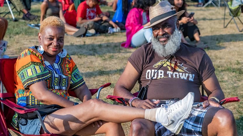 an african american couple sit in folding chairs in a sunny field. the woman has a foot on the man's lap.
