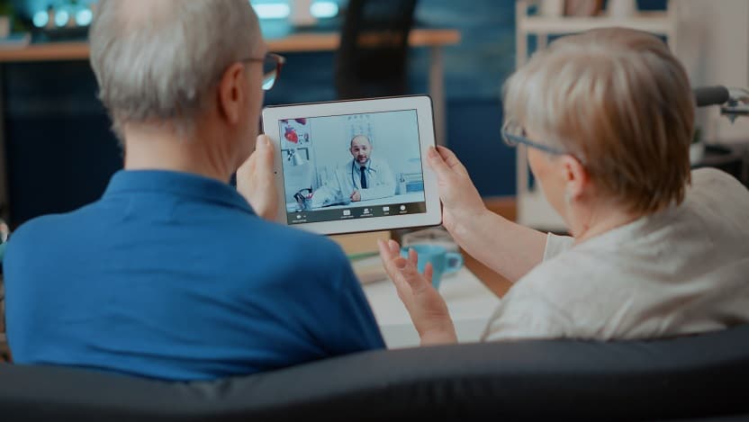 An elderly couple holds an iPad showing a video call with a doctor