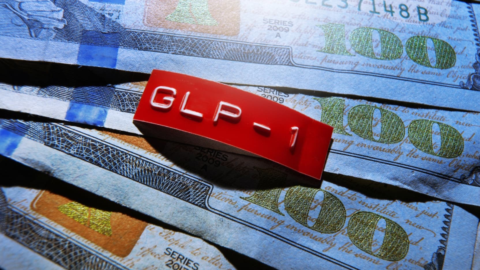 A red "GLP-1" label rests on a spread of $100 bills.