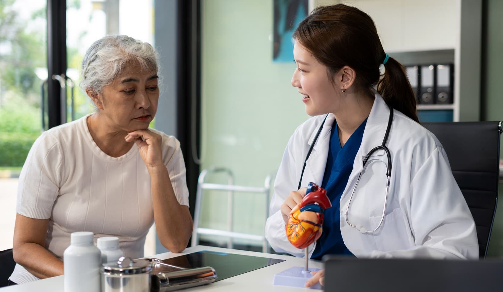 A doctor and patient converse at a table with an anatomical model of a heart on it.