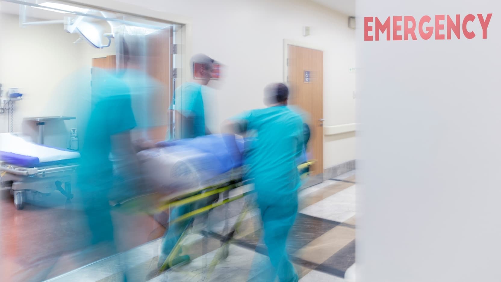 A blurred image of three nurses in blue scrubs rushing a patient on a stretcher past an 'Emergency' sign