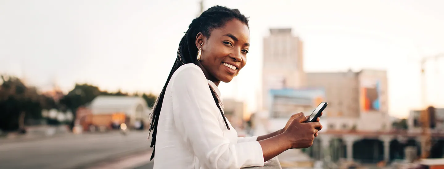 A woman in a city standing while holding her phone and smiling at the camera