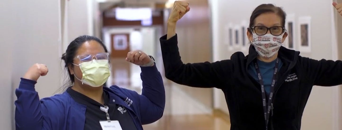 Two female nurses wearing masks smile at the camera and flex their muscles in a power pose