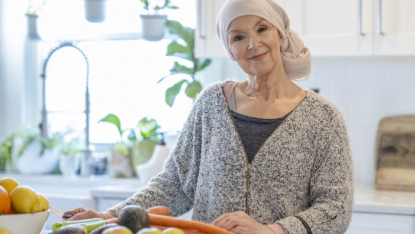 An elderly female cancer patient stands at a kitchen counter with some vegetables in front of her