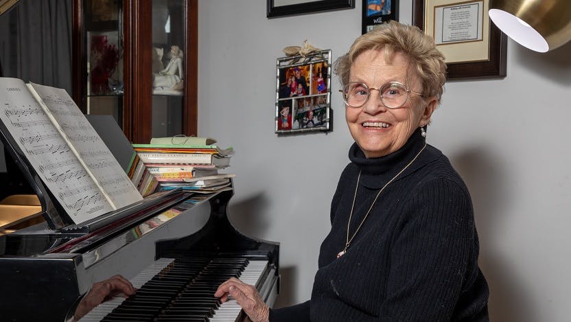 Marcia Pavich, an older white woman with short hair, sits at a piano smiling with a wall of picture frames behind her